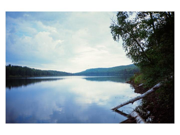 A calm lake reflects the sky and surrounding forest under a cloudy sky.