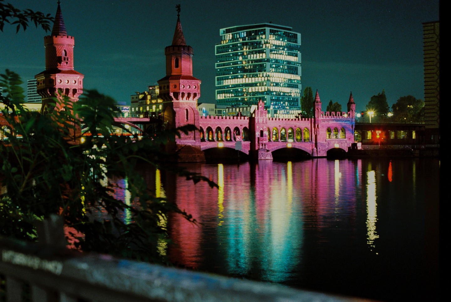 A bridge with two towers is illuminated in pink and purple light, with a modern building and its reflection in the water at night.