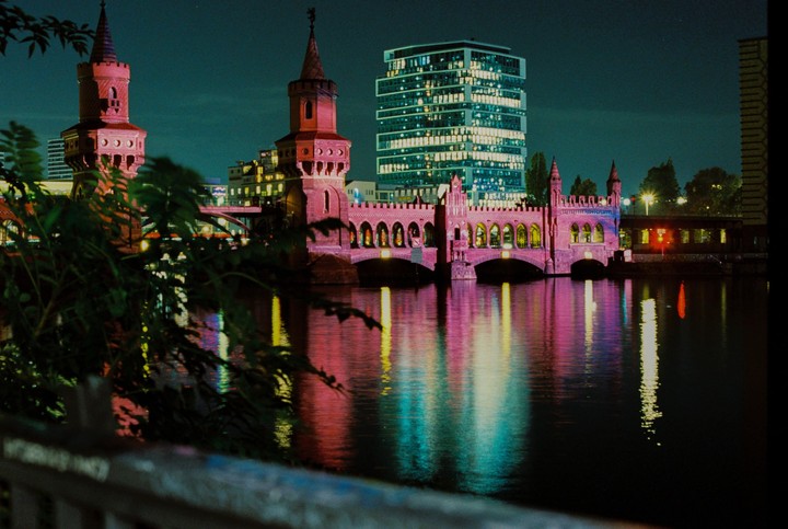 A bridge with two towers is illuminated in pink and purple light, with a modern building and its reflection in the water at night.