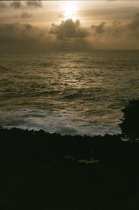 The sun shines through clouds over a rocky coastline and ocean waves.