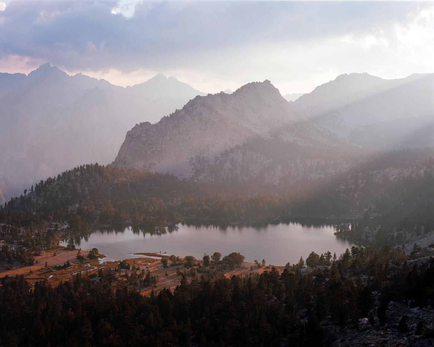 Sunlight streams through clouds over a mountain lake surrounded by pine trees and rocky terrain.