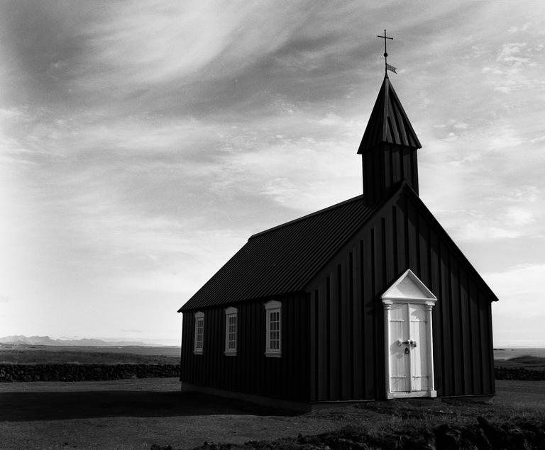 A black church with a white door and steeple stands in a field under a cloudy sky.