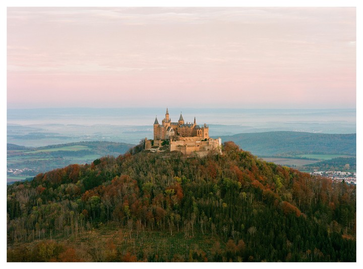 A castle with multiple turrets and spires sits atop a forested hill under a pale pink sky.