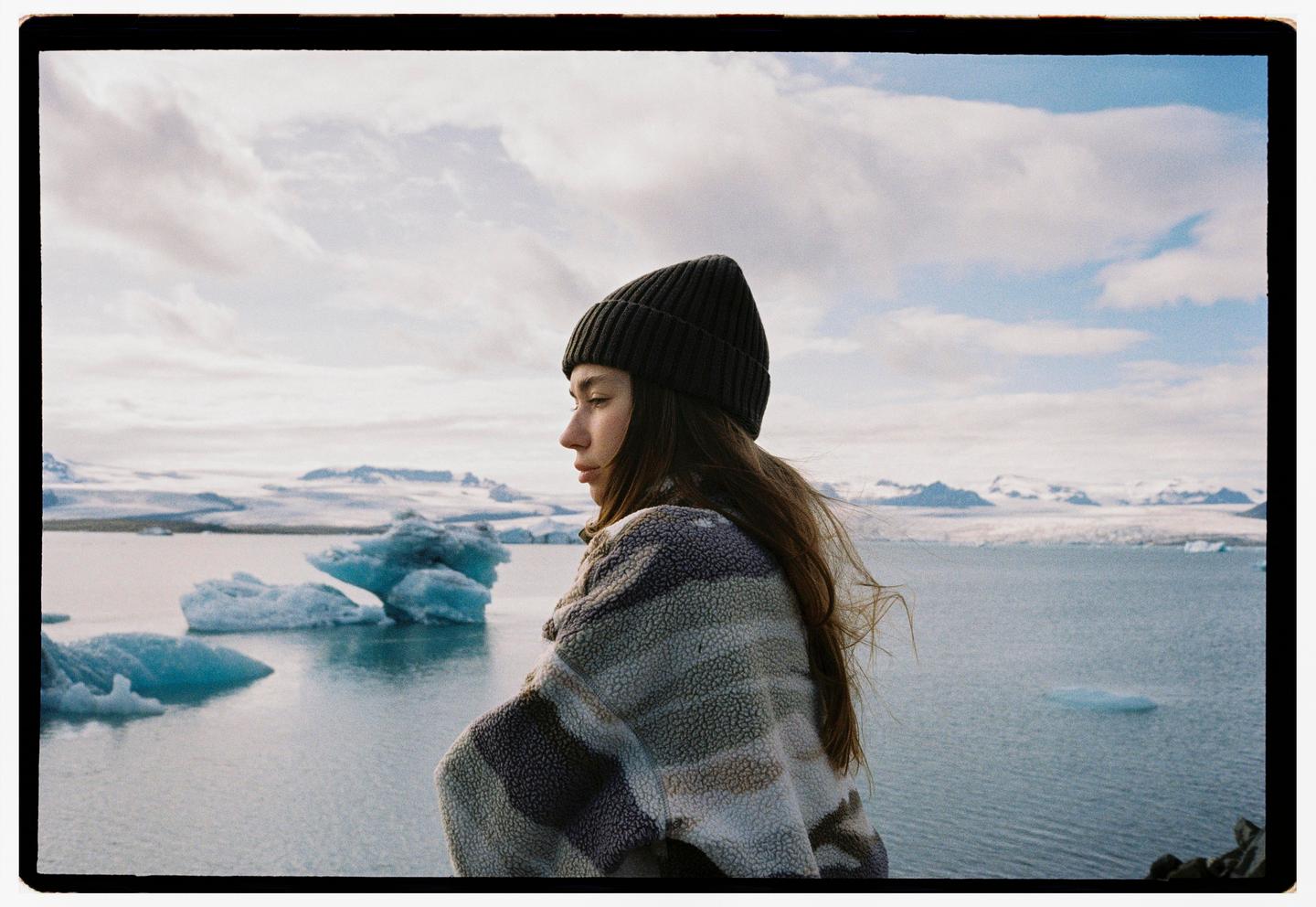 A woman wearing a beanie and a patterned jacket looks out over a glacial lagoon with icebergs and snow-covered mountains in the distance.