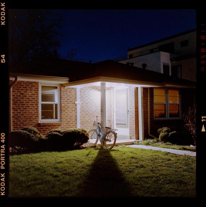 A bicycle is parked on a lawn in front of a brick house with a lit porch at night.