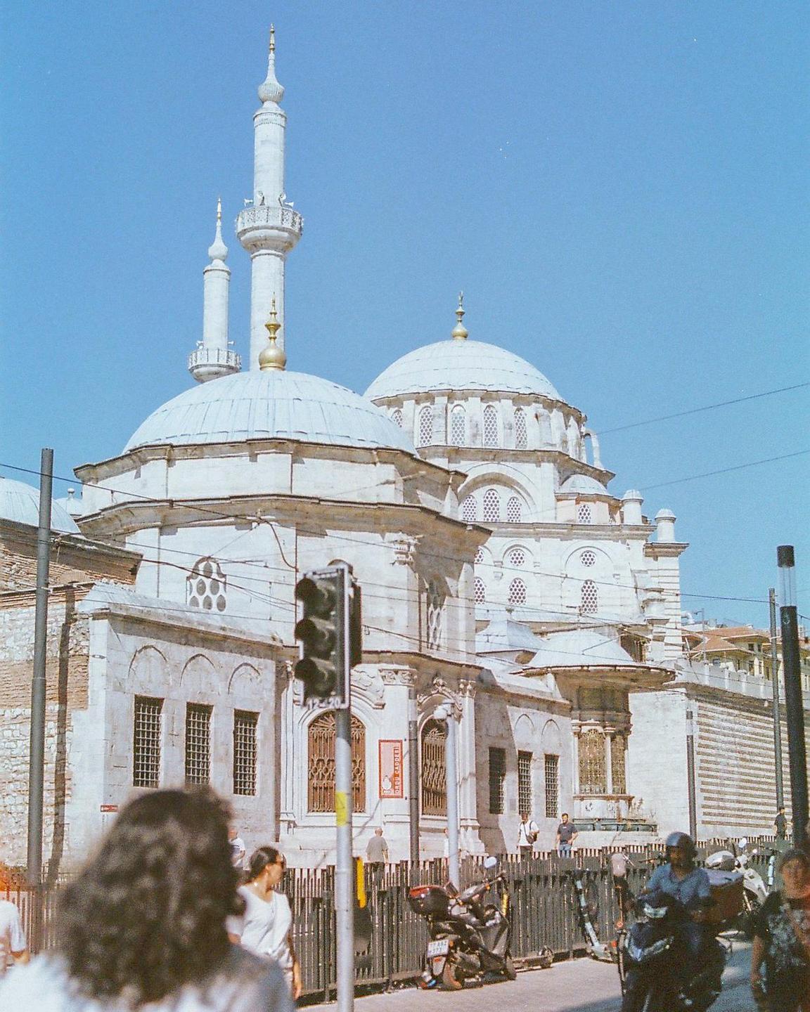 A large white mosque with multiple domes and minarets stands prominently against a clear blue sky, with a street and people in the foreground.