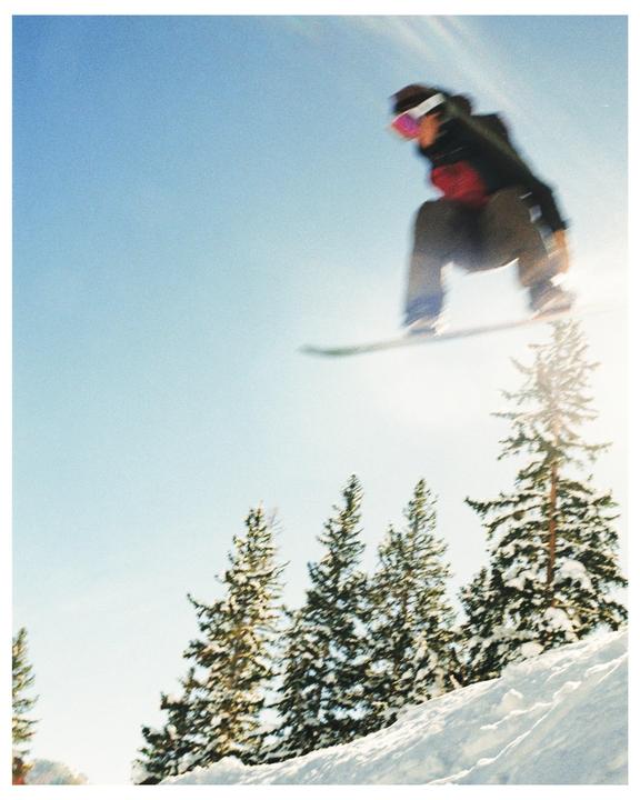 A snowboarder jumps over snow-covered pine trees on a sunny day.