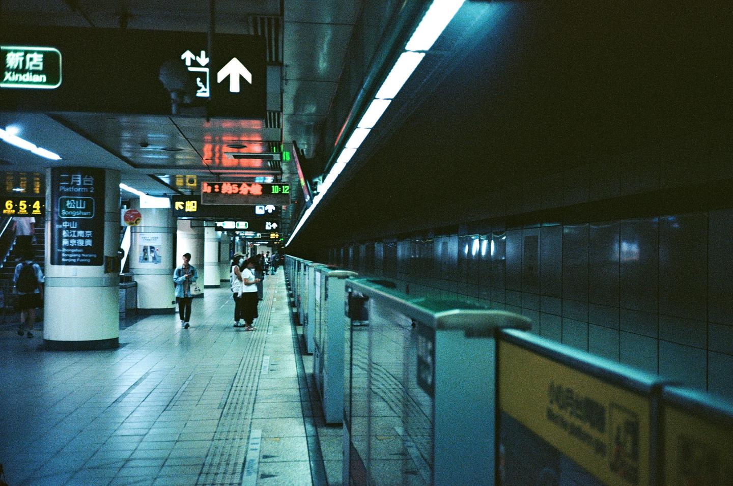 People wait on a subway platform with bright overhead lights and signs in an underground station.