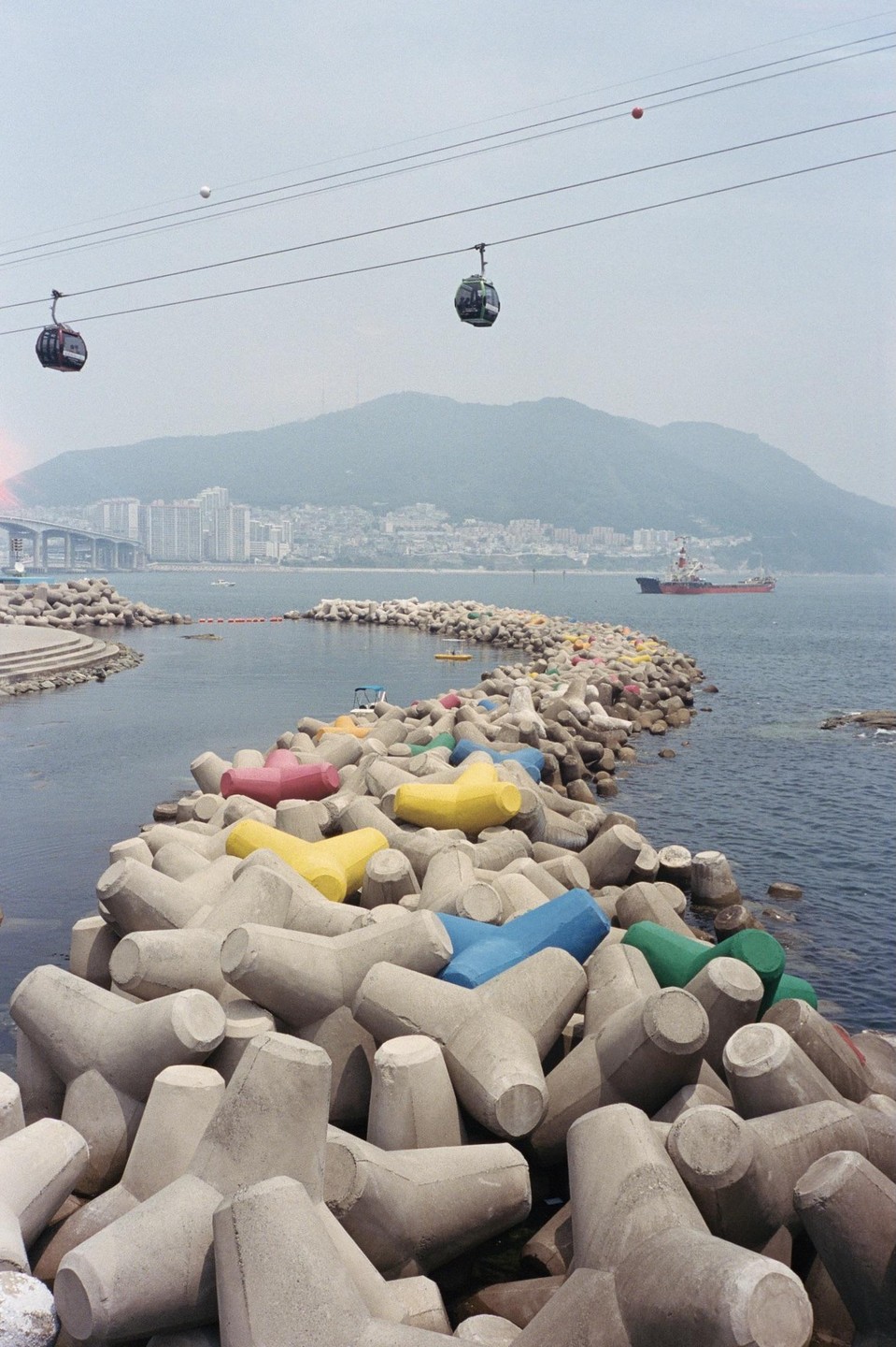 Two cable cars pass over a breakwater made of concrete tetrapods, some painted in bright colors, with a city and mountain in the background.
