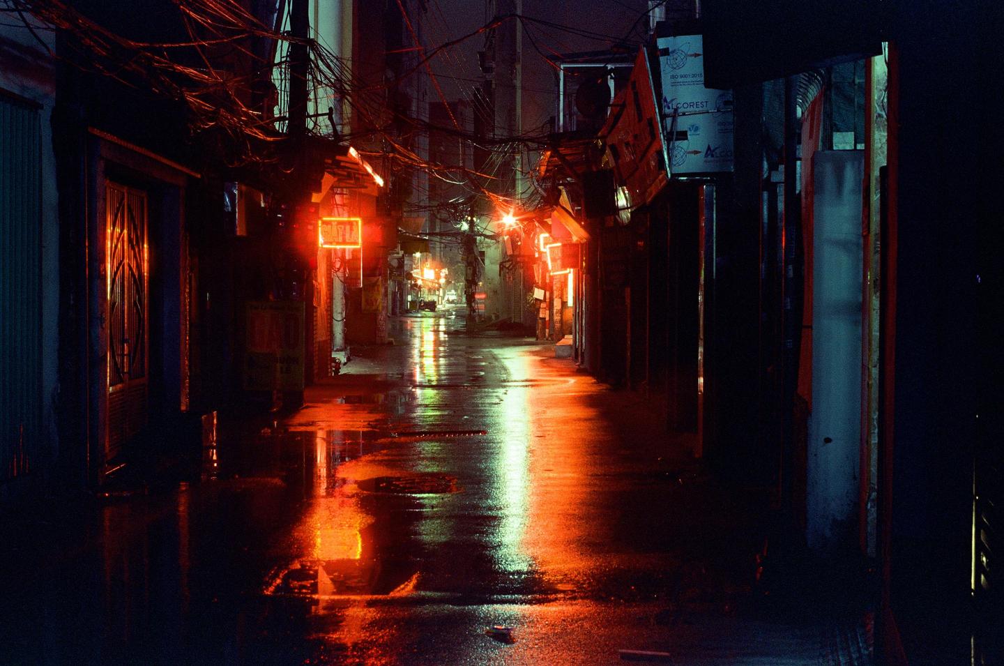 A narrow street is illuminated by neon signs and reflected in the wet pavement at night.