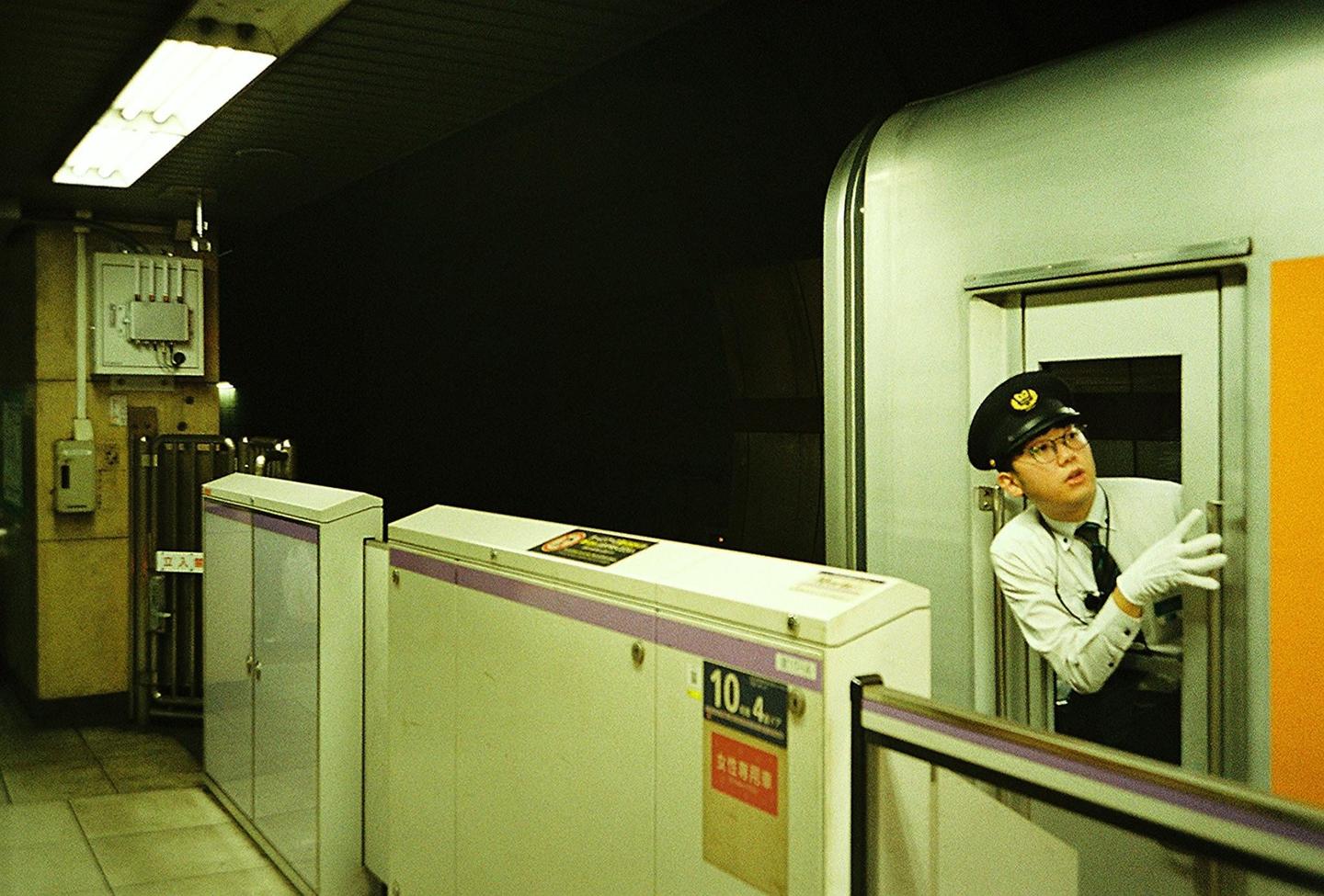 A man in a uniform and hat looks out from a train door at a station platform at night.