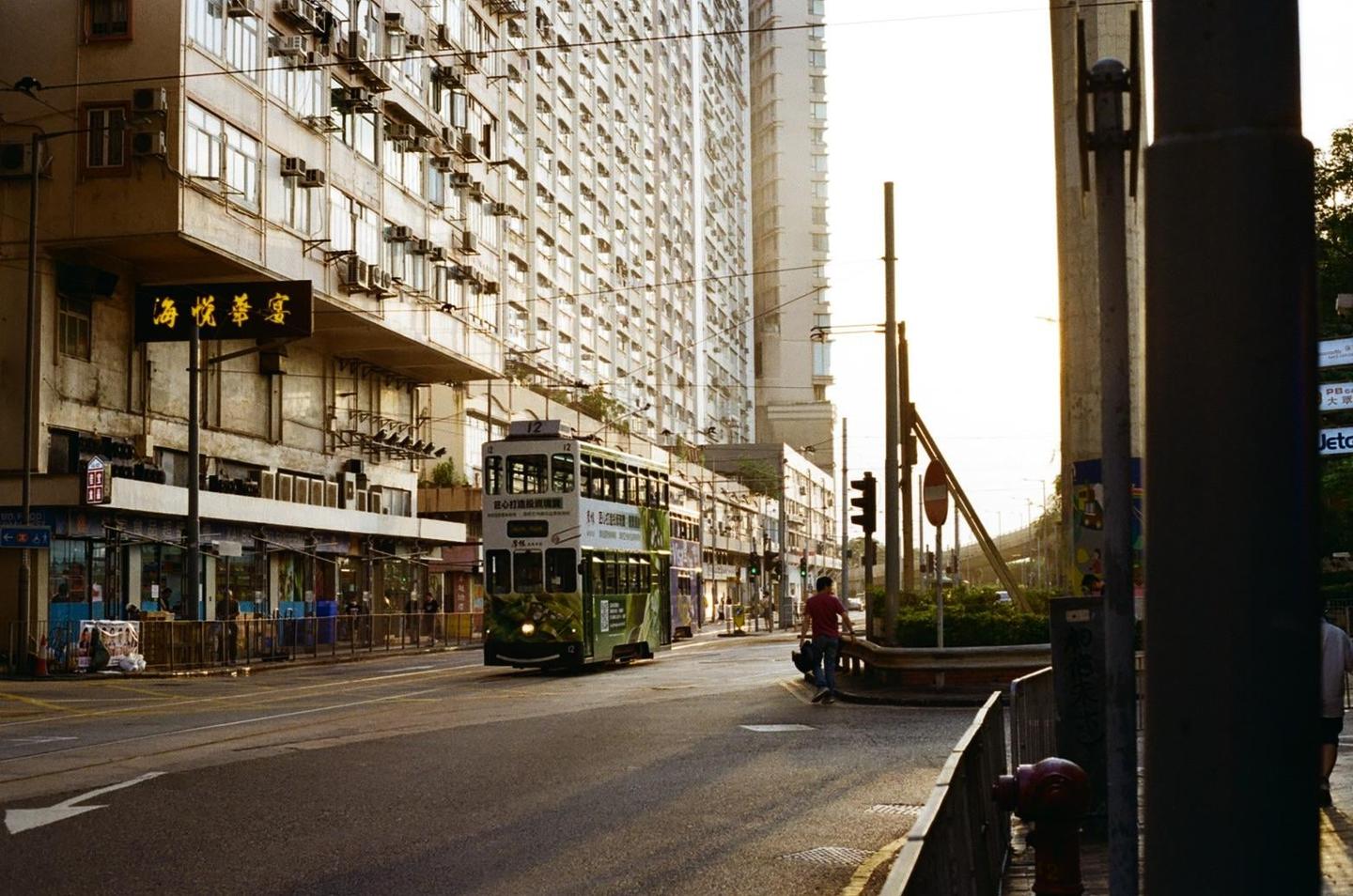 A green double-decker tram travels down a city street lined with tall buildings and a man walks on the sidewalk.