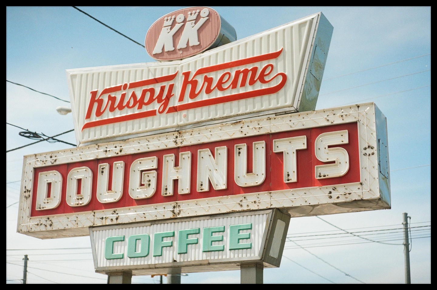 A weathered Krispy Kreme sign advertising doughnuts and coffee stands against a clear blue sky.