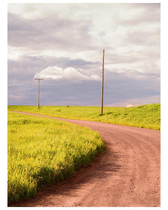 A dirt road curves through a vibrant green field under a cloudy sky with telephone poles and wires.