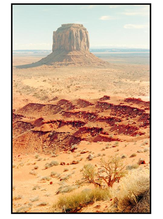 A vast desert landscape features a large mesa under a pale sky, with smaller red rock formations and sparse vegetation in the foreground.
