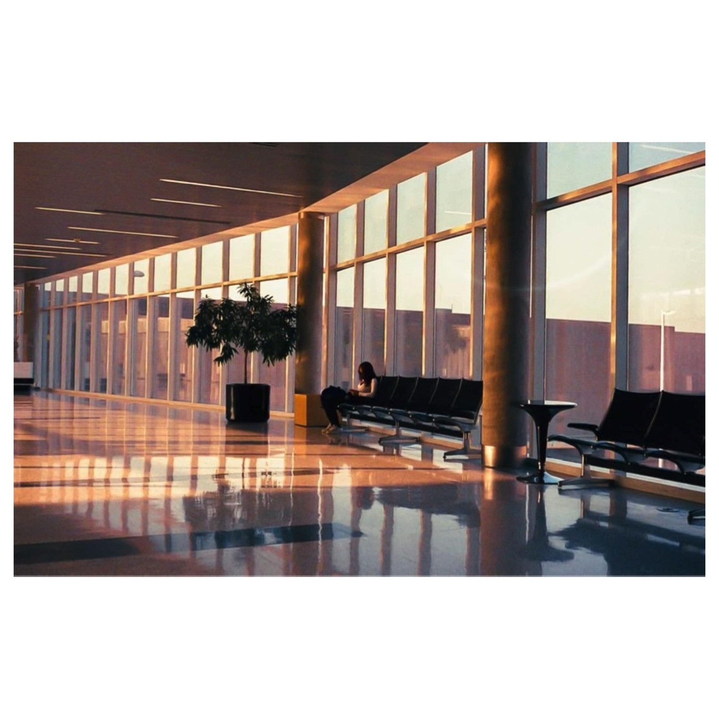 A woman sits on a bench in a large, sunlit hall with many windows.