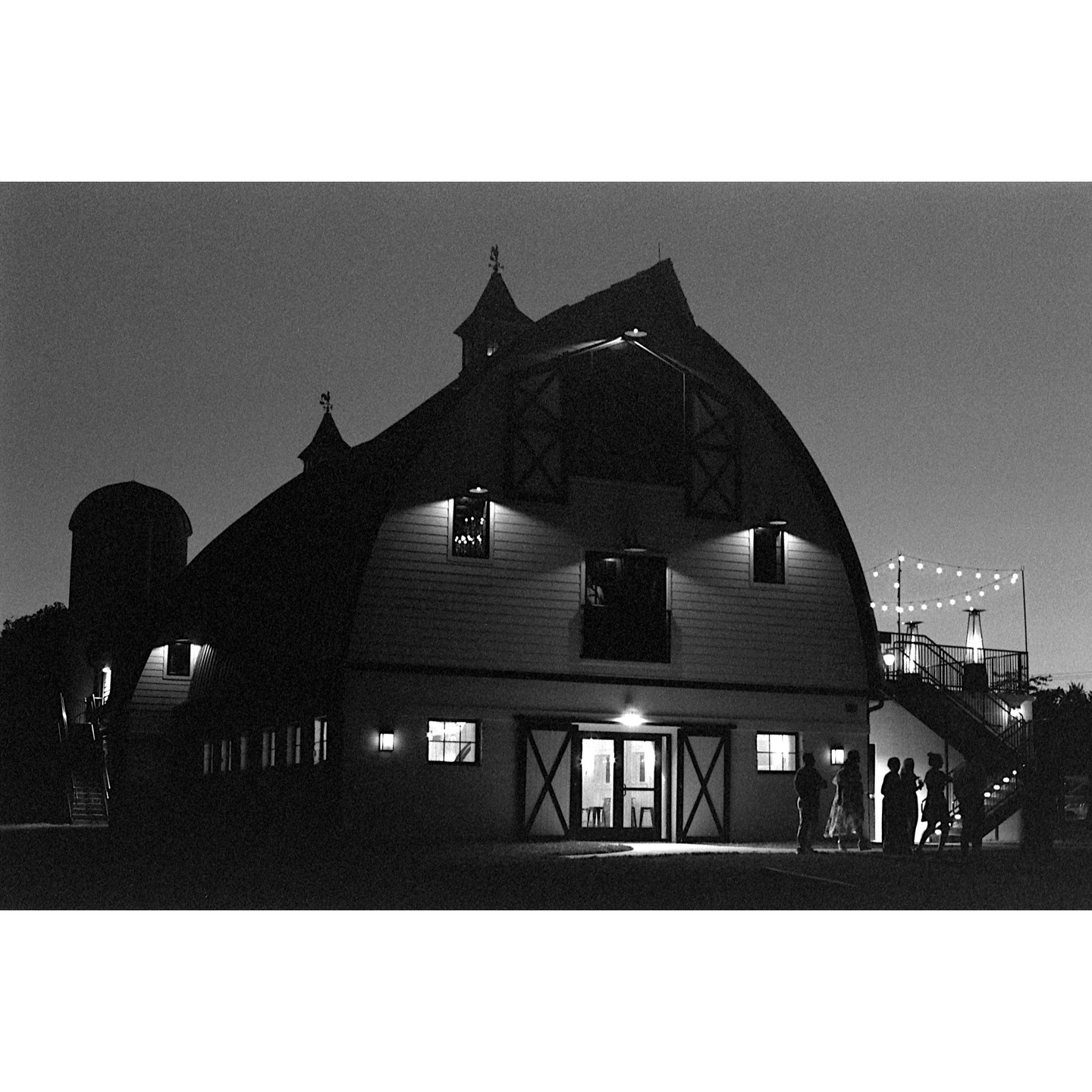 A large barn with a silo and string lights is illuminated at night, with several people standing near the entrance.