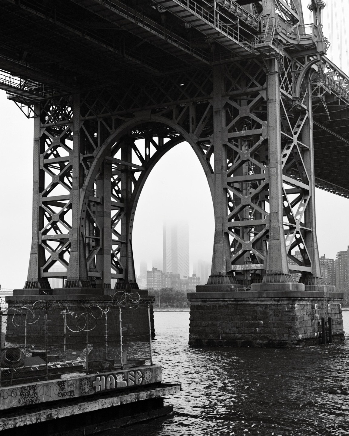 A black and white photograph shows the underside of a large bridge with an arched support structure, with a city skyline visible through a misty background.