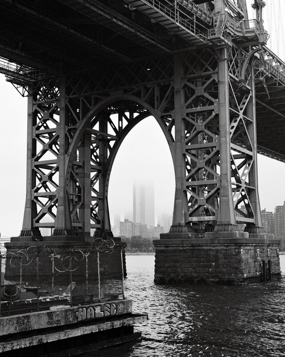A black and white photograph shows the underside of a large bridge with an arched support structure, with a city skyline visible through a misty background.