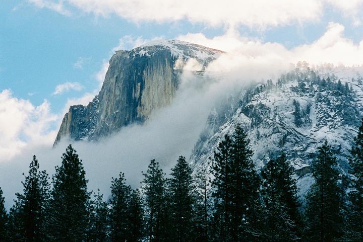 Clouds partially obscure a snow-covered Half Dome and surrounding pine trees.