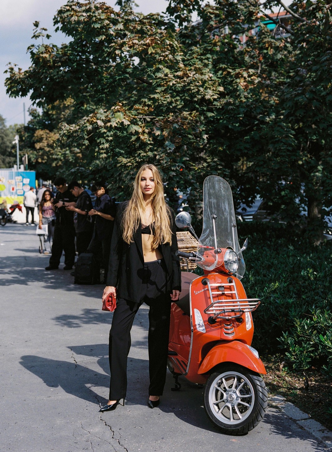 A woman in a black suit stands next to an orange scooter on a paved street.
