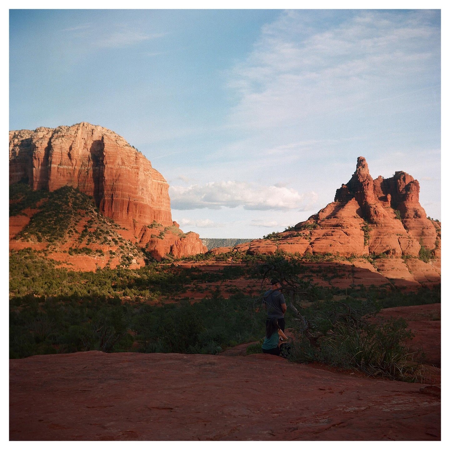 A man and a woman stand on a red rock formation overlooking a valley with large red rock mountains under a blue sky.