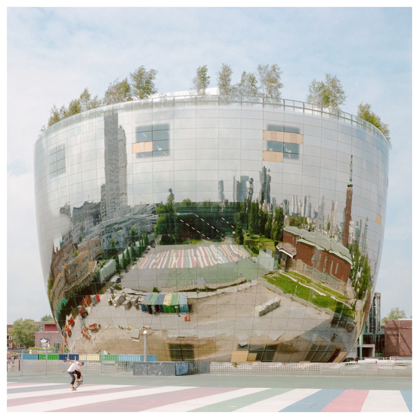 A person on a skateboard is in front of a large, round, reflective building with trees on its roof.