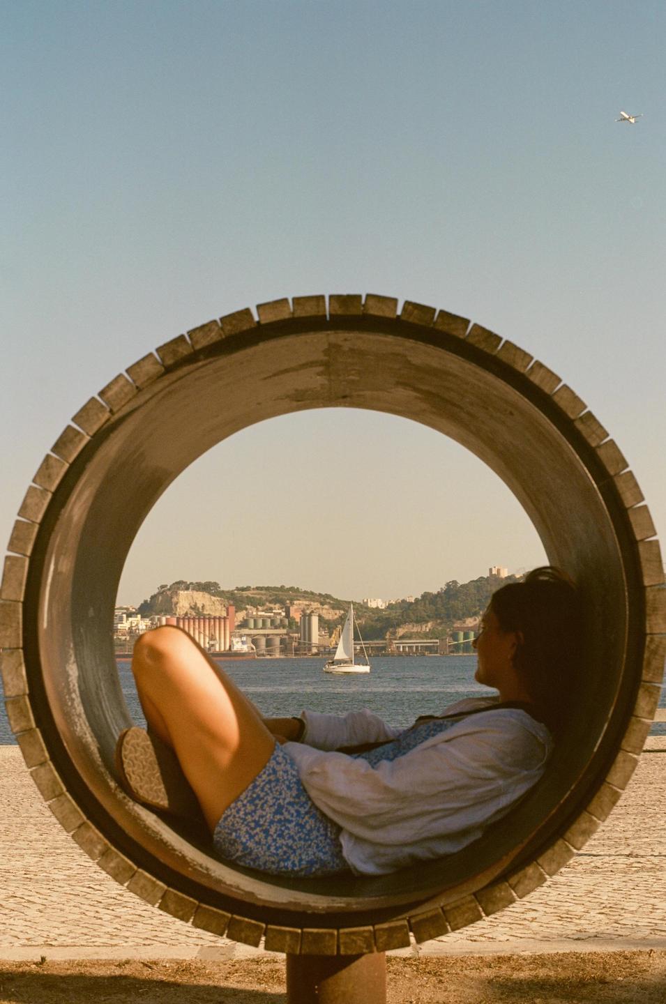 A woman is relaxing inside a large wooden ring, looking out at a sailboat on the water with a city and hills in the background.