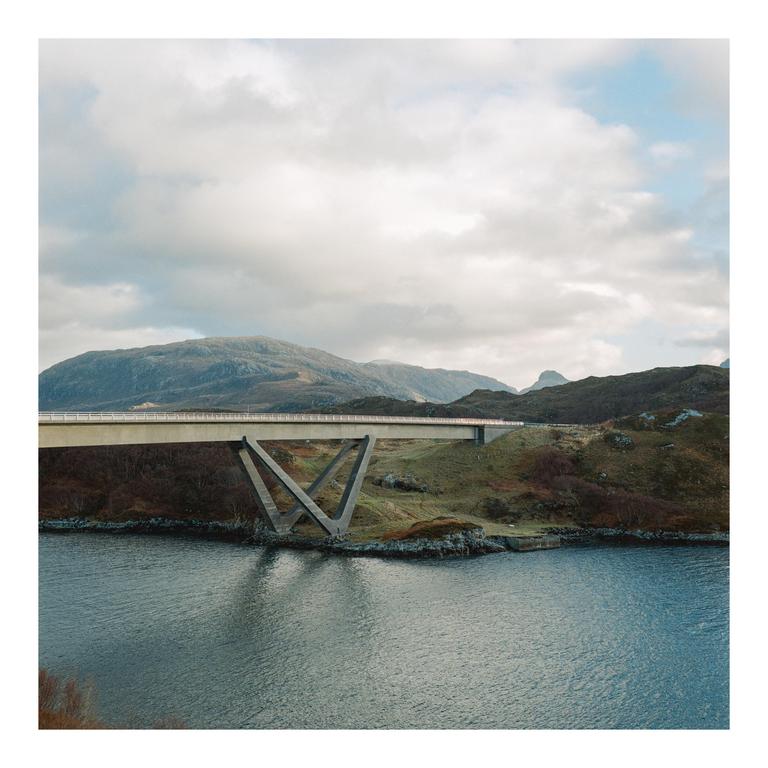 A concrete bridge with a distinctive V-shaped support spans a body of water, connecting two hilly landmasses under a cloudy sky.