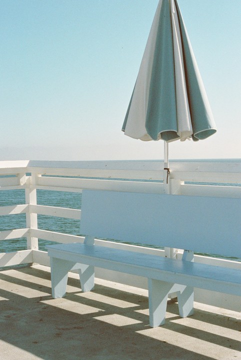 A blue and white striped umbrella stands next to a white bench on a pier overlooking the ocean.