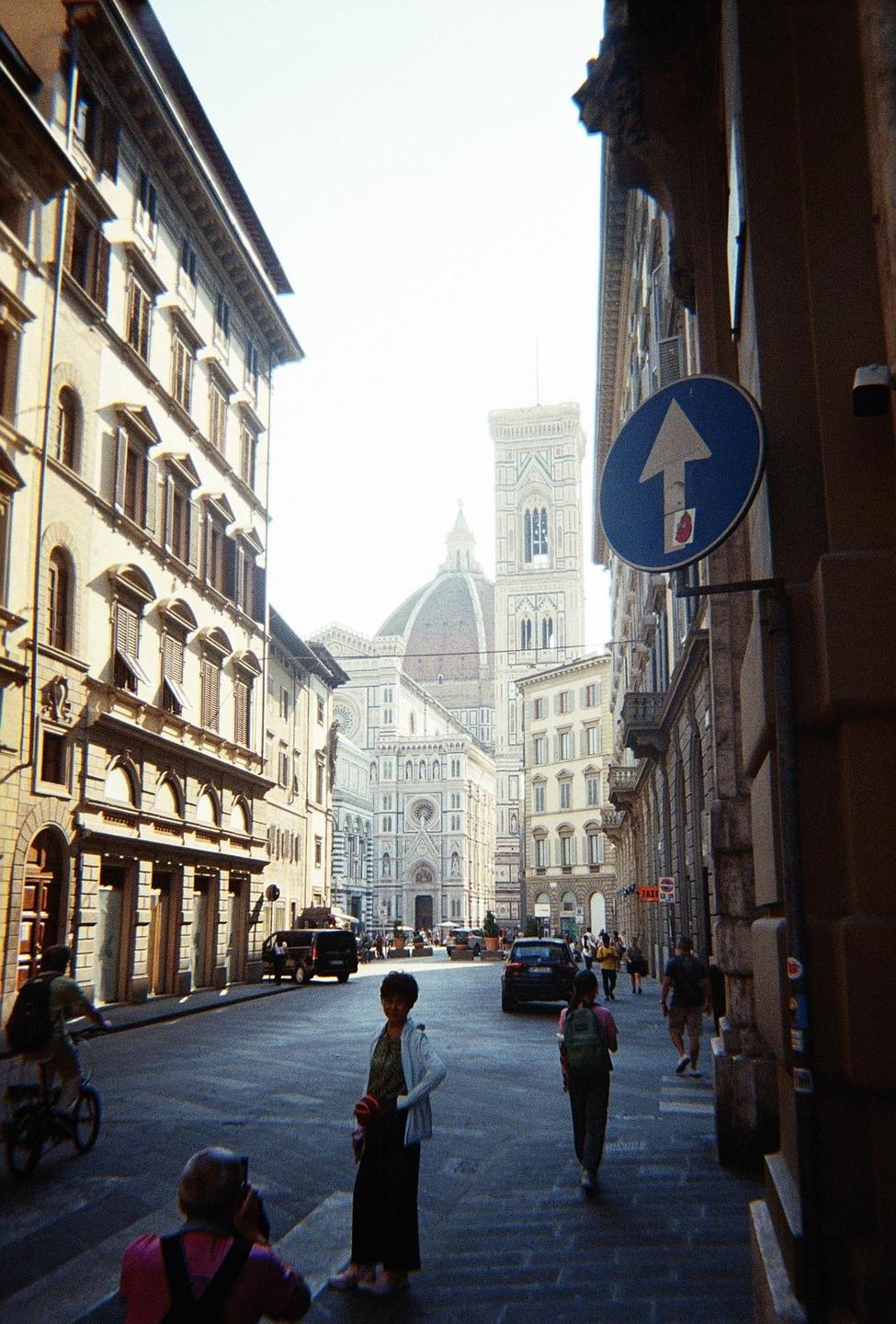 A street in Florence leads to the Duomo, with people walking and a blue arrow sign on the right.