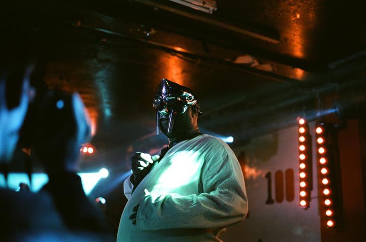 A man wearing a metal mask and a light-colored shirt stands on a stage with colorful lights.