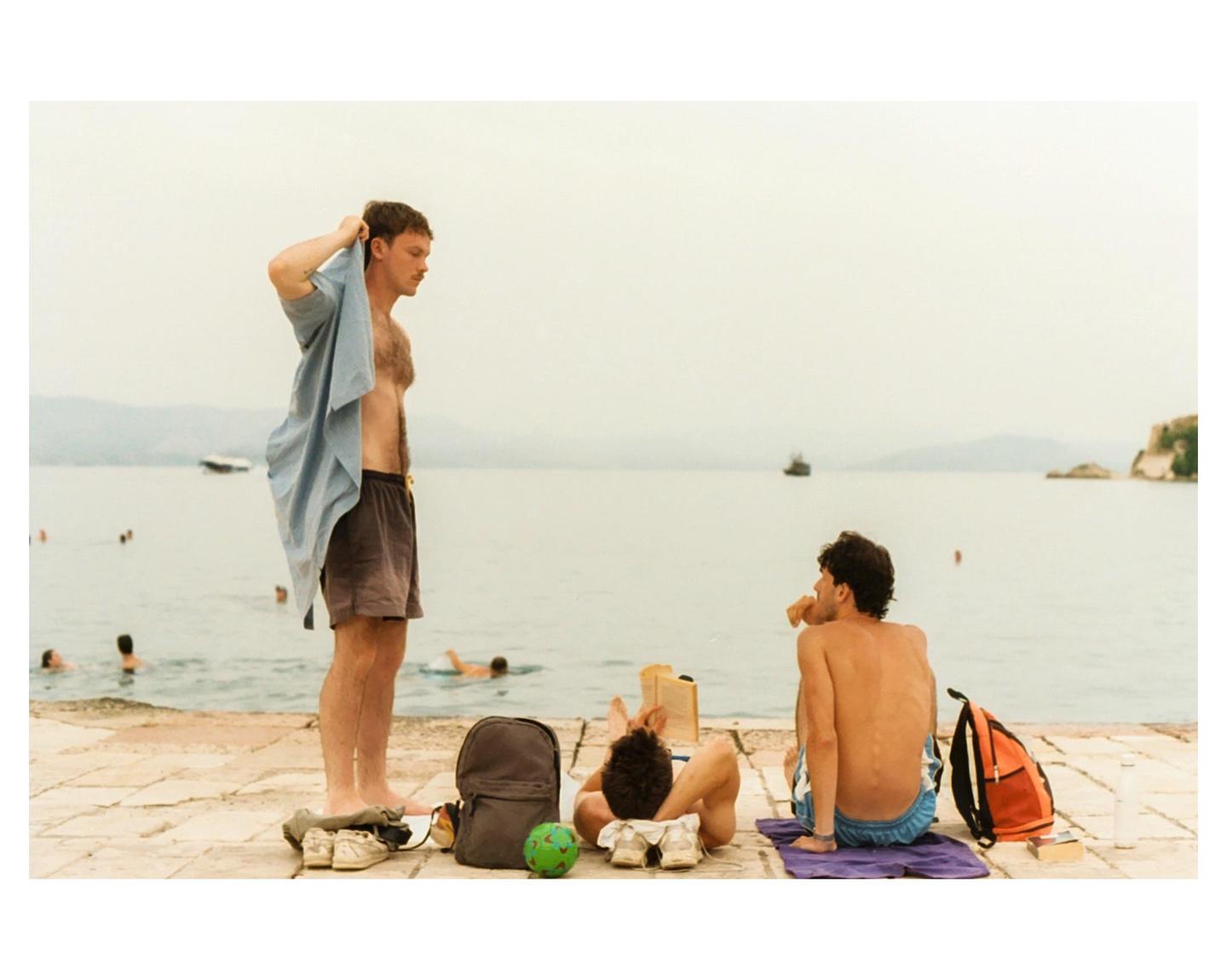 Three men relax by the water, one standing and two sitting, with a boat and land visible in the distance.
