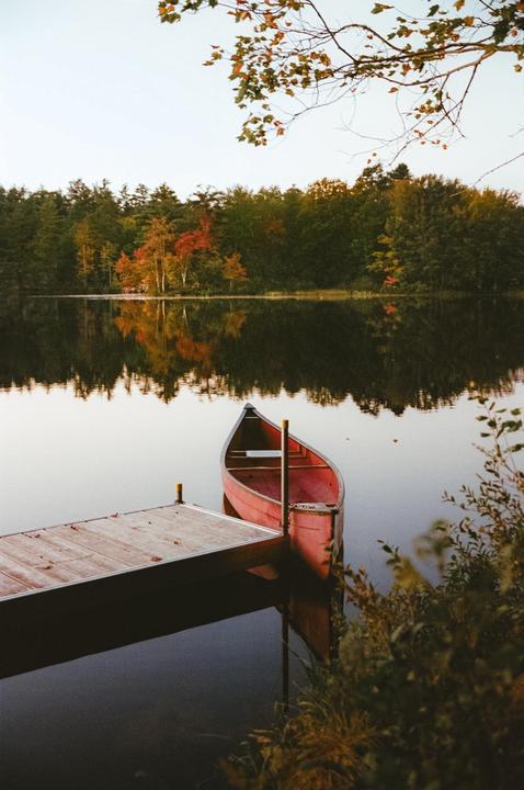 A red canoe is docked next to a wooden pier on a calm lake surrounded by autumn trees.