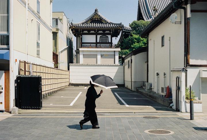 A person holding an umbrella walks past a parking lot with a traditional Japanese building in the background.