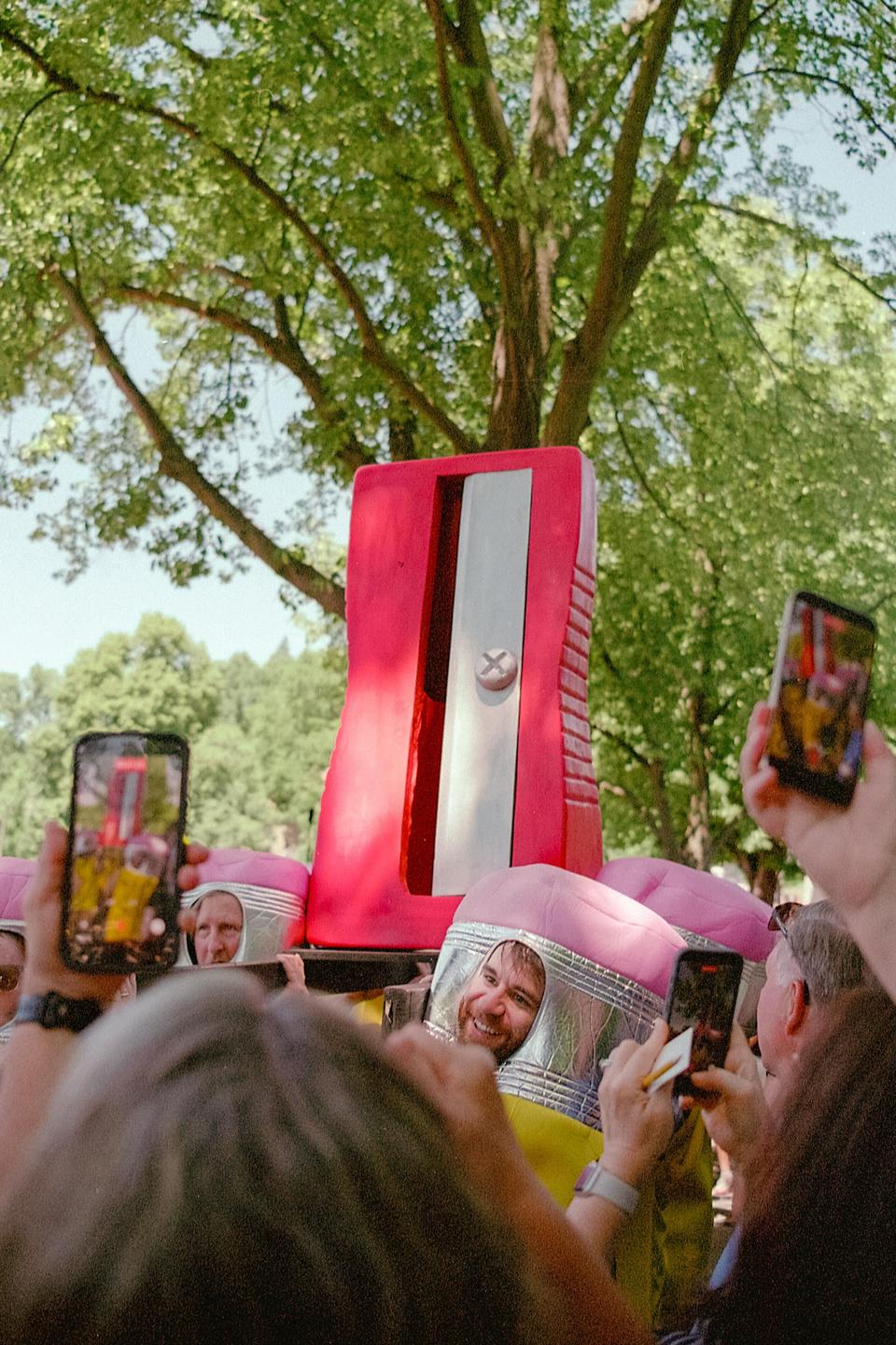 A man in a pencil costume smiles at the camera while people around him take photos with their phones, with a giant pencil sharpener in the background.