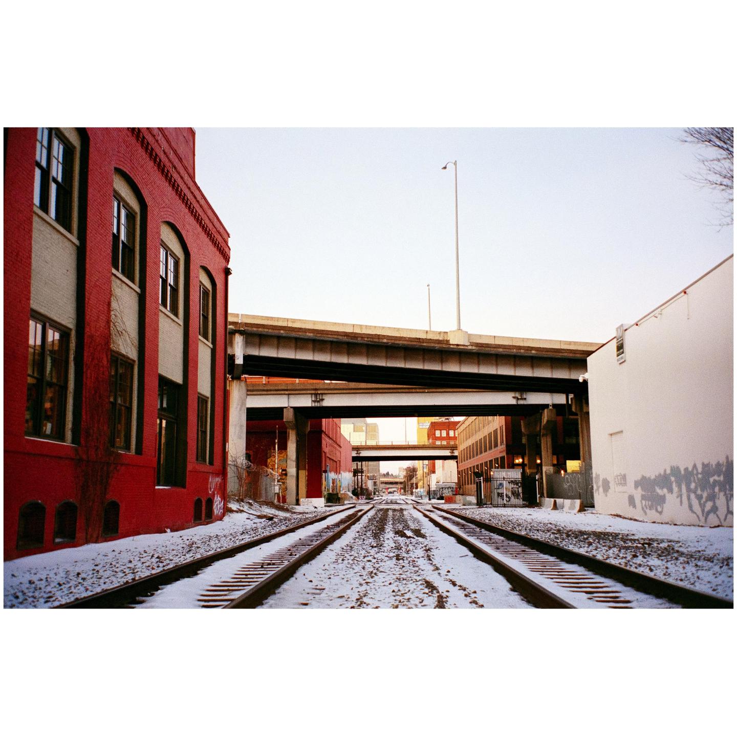 Snow-covered train tracks lead toward a city under an overpass, with a red brick building on the left and a white building on the right.