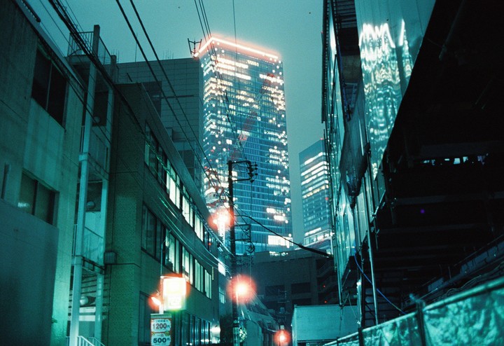 A street at night is lined with buildings and power lines, with a tall, illuminated skyscraper in the background.