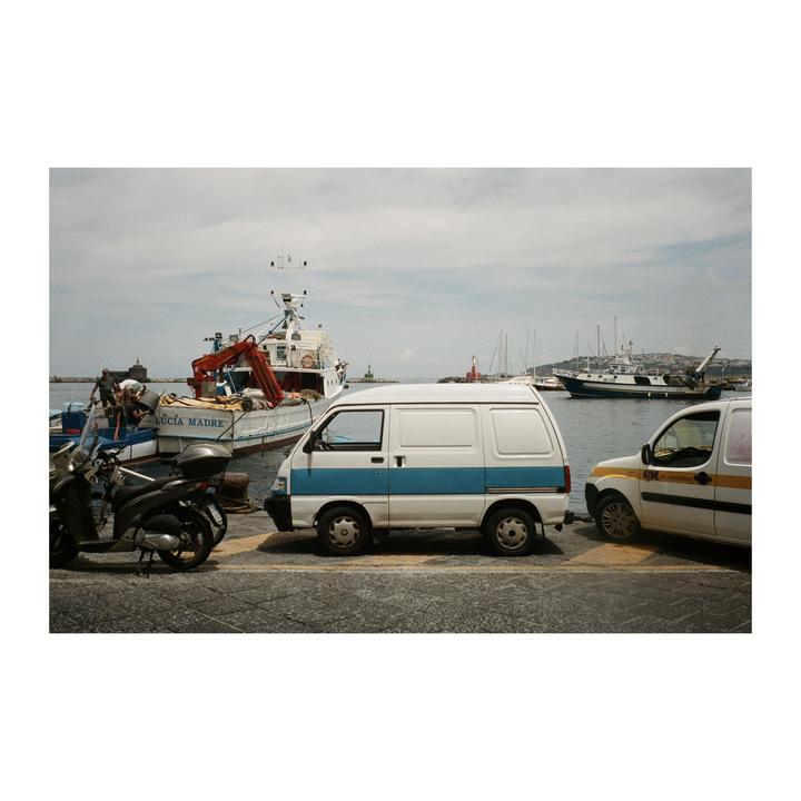 Two vans and a scooter are parked on a paved surface next to a harbor with several boats.