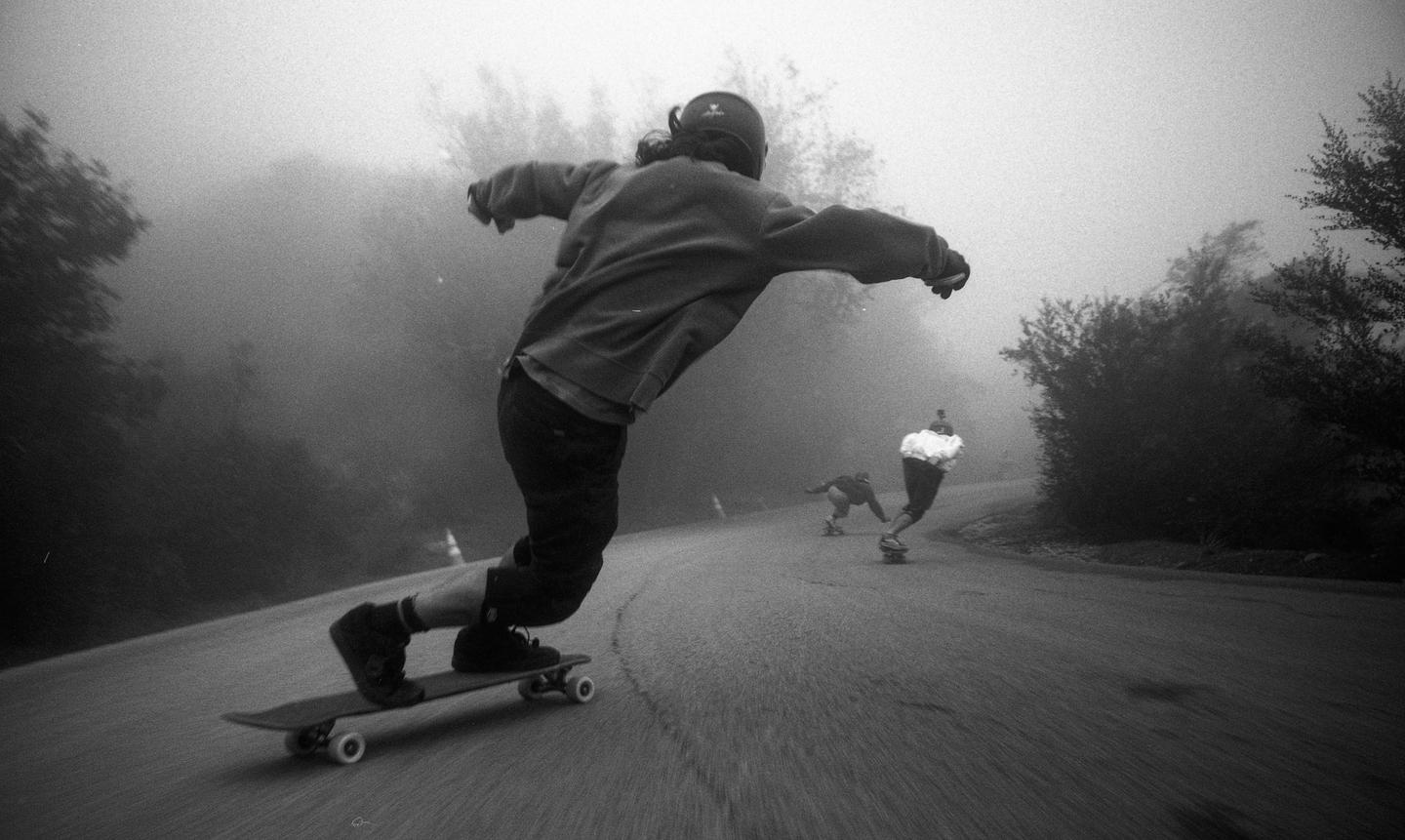 Three people are skateboarding down a winding road in the fog.