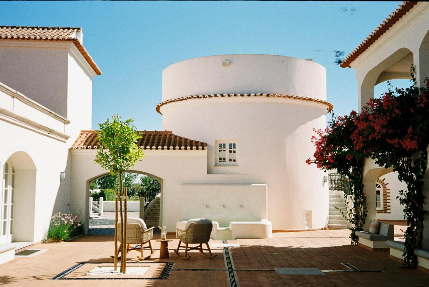 A courtyard with white buildings, terracotta roofs, and a small tree in the center is shown under a clear blue sky.
