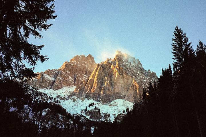 The last light of day illuminates the peaks of a snow-covered mountain range, framed by dark evergreen trees.