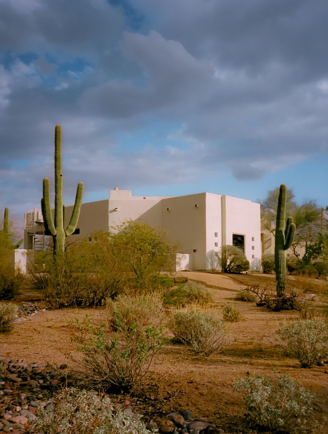 A white building with a spiral staircase is surrounded by desert plants and cacti under a cloudy sky.