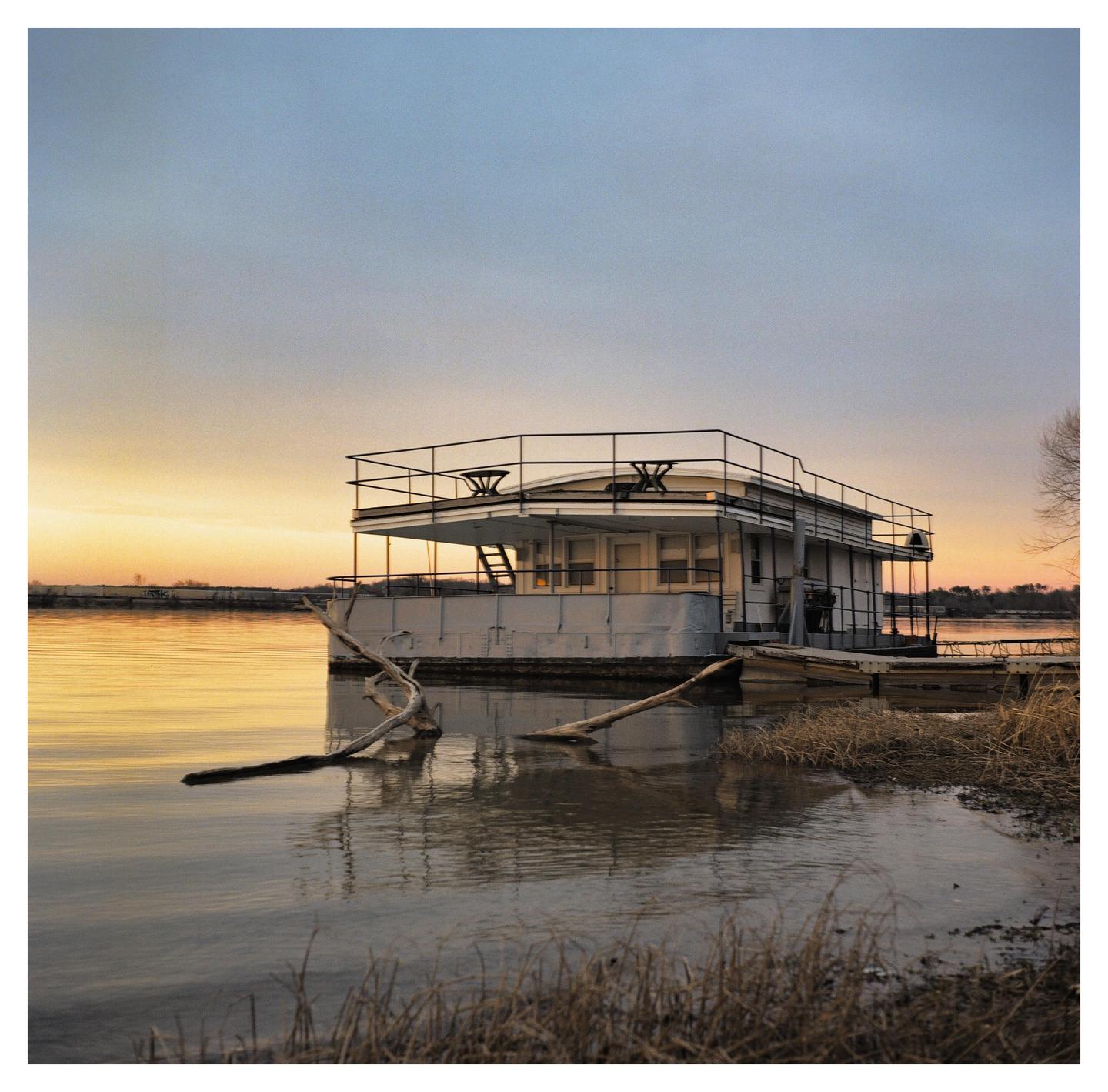 A houseboat is moored on a river at sunset, with bare branches and dry grass in the foreground.