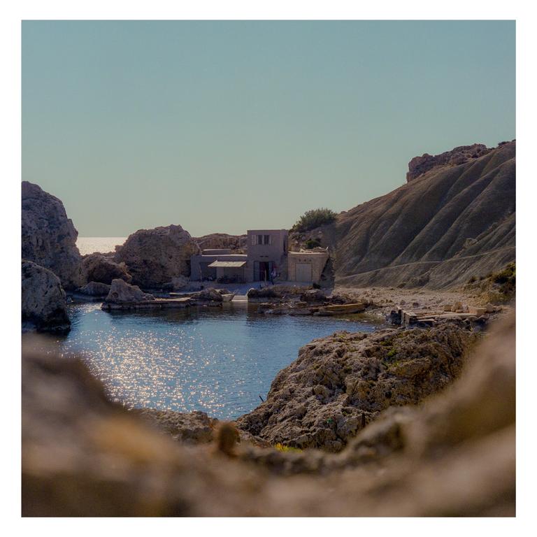 A small building is nestled among rocky cliffs beside a calm body of water with the sea visible in the distance.