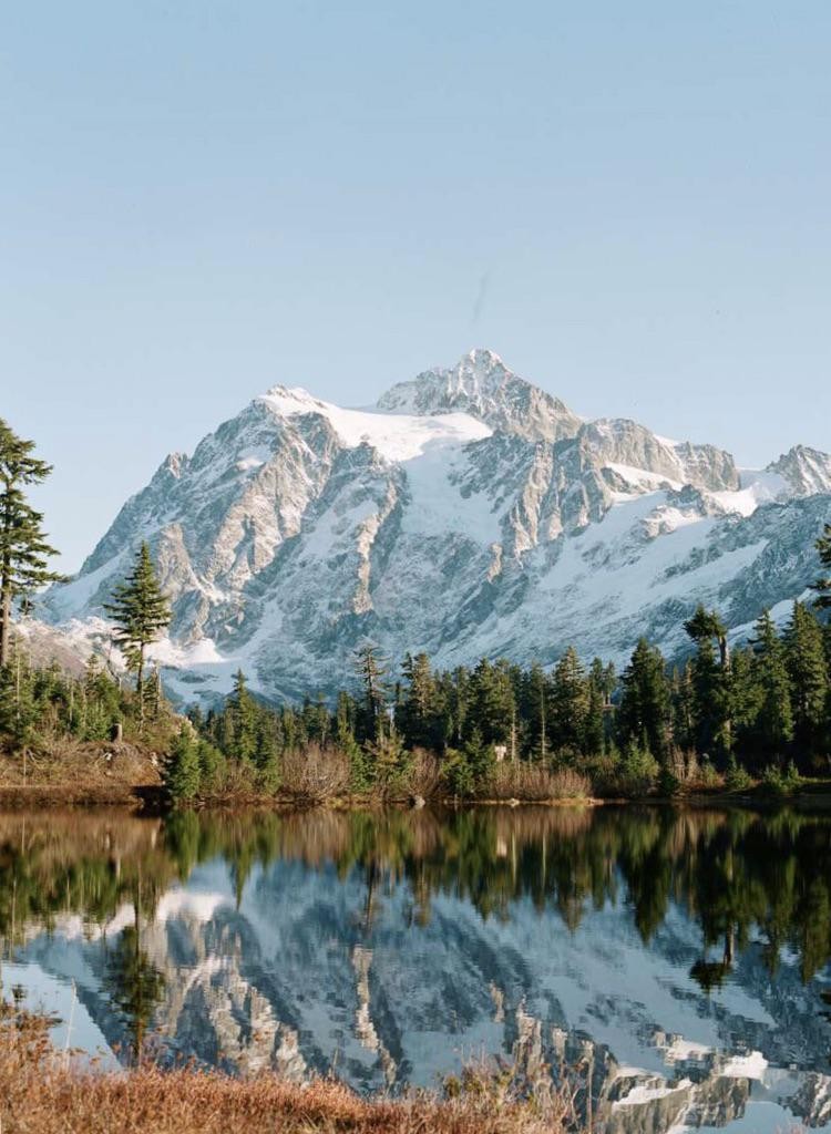 A snow-capped mountain is reflected in a still lake surrounded by a forest.