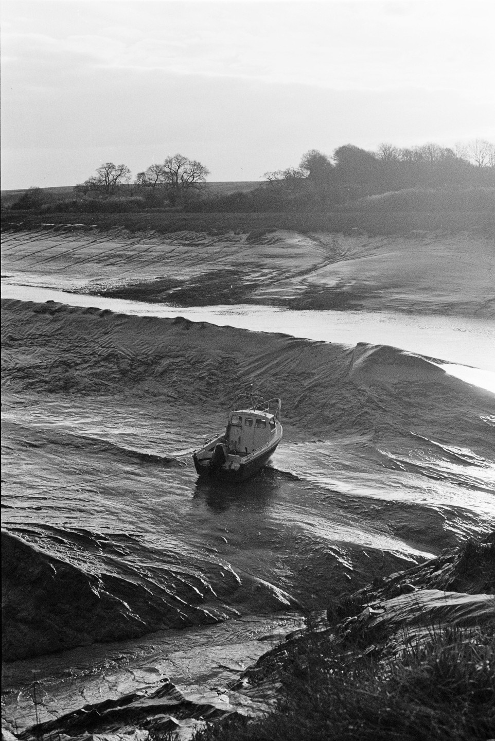 A small boat is beached on a muddy riverbank with trees on the distant shore under a bright sky.