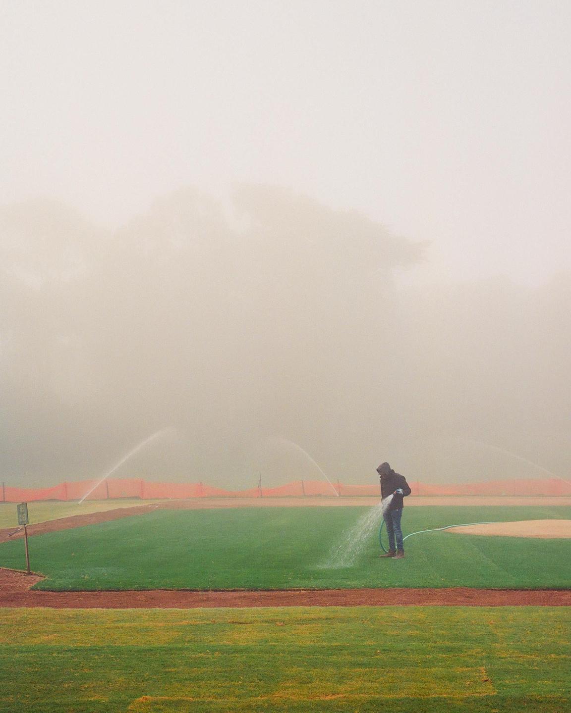 A man waters a baseball field on a foggy morning.