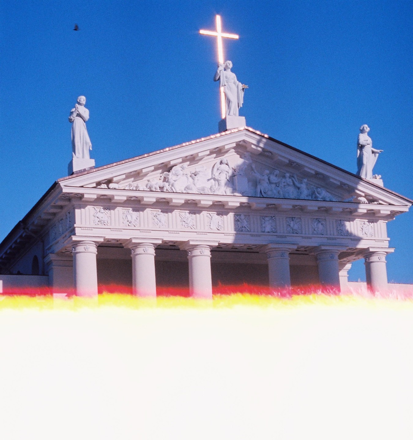 A white neoclassical building with statues and a glowing cross on its roof stands against a clear blue sky, with a bright light leak across the bottom.