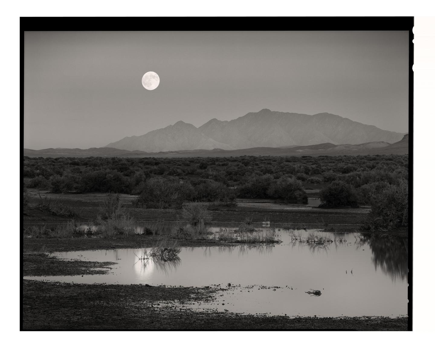 A full moon rises over a desert landscape with mountains in the distance and a body of water reflecting the moon in the foreground.